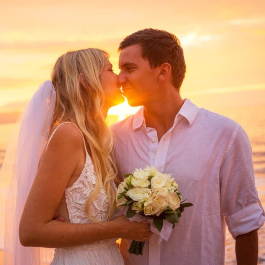 A couple shares a kiss at sunset, with the bride holding a bouquet of roses.