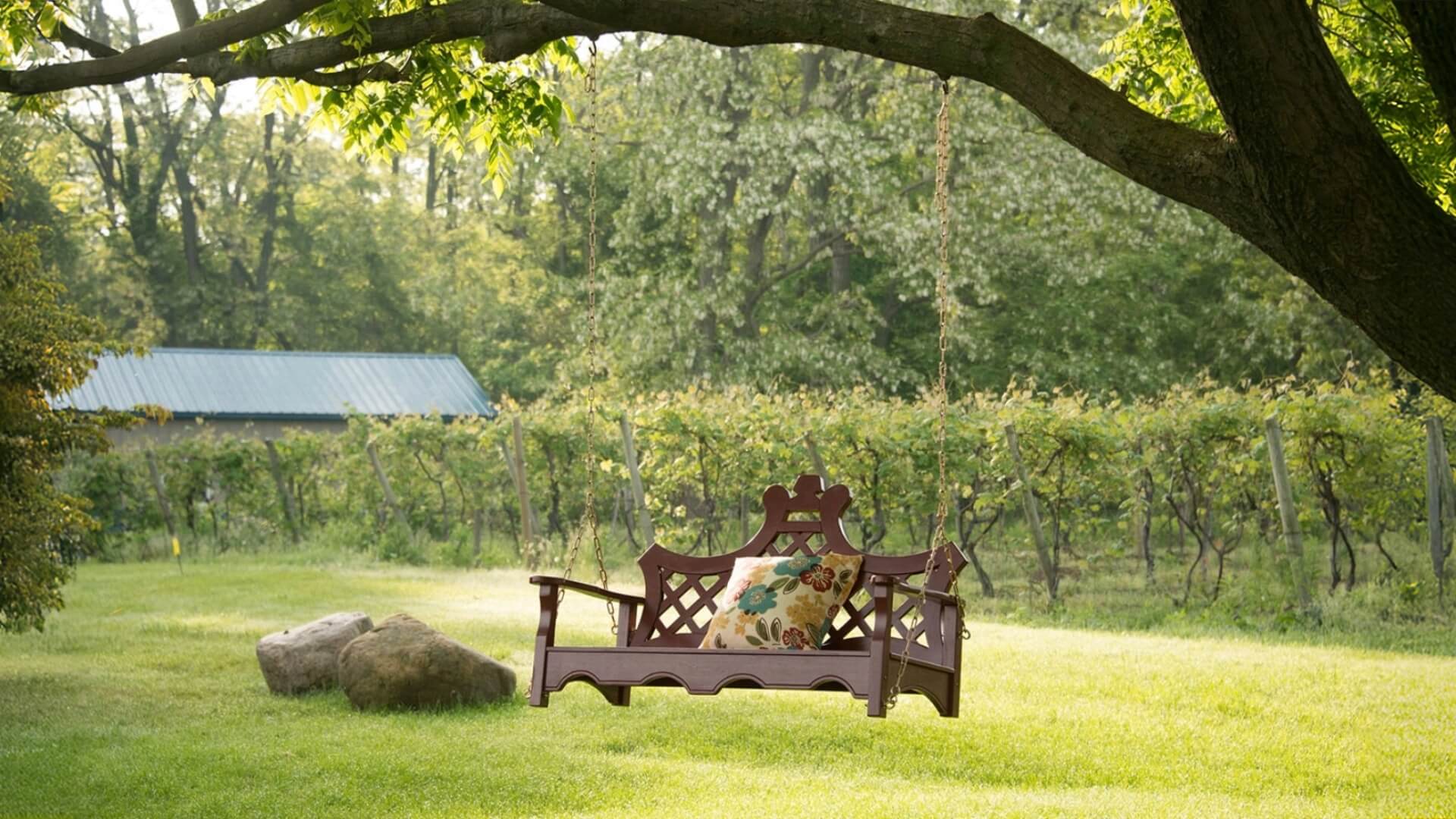 A wooden bench with a floral cushion under a tree in a grassy area near a vineyard.