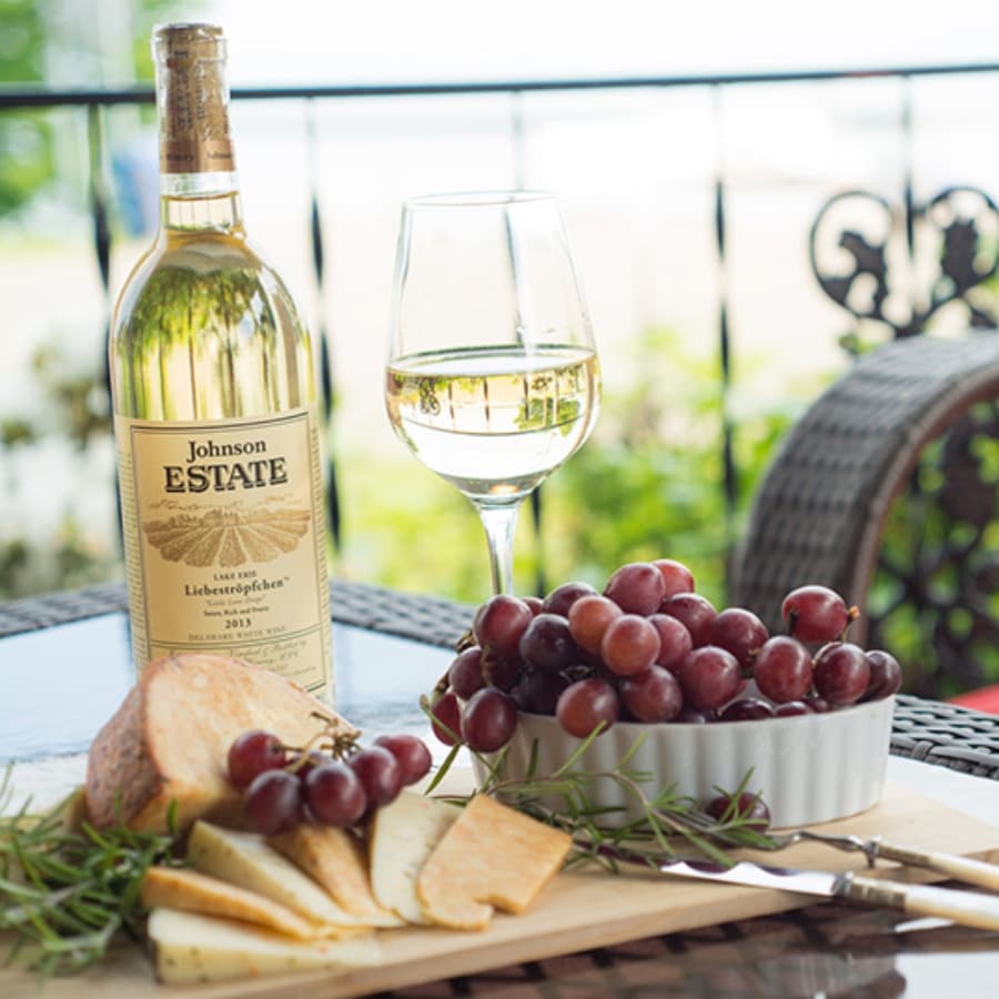 A bottle of white wine, a glass, and a plate of cheese, grapes, and bread on a table with a scenic background.