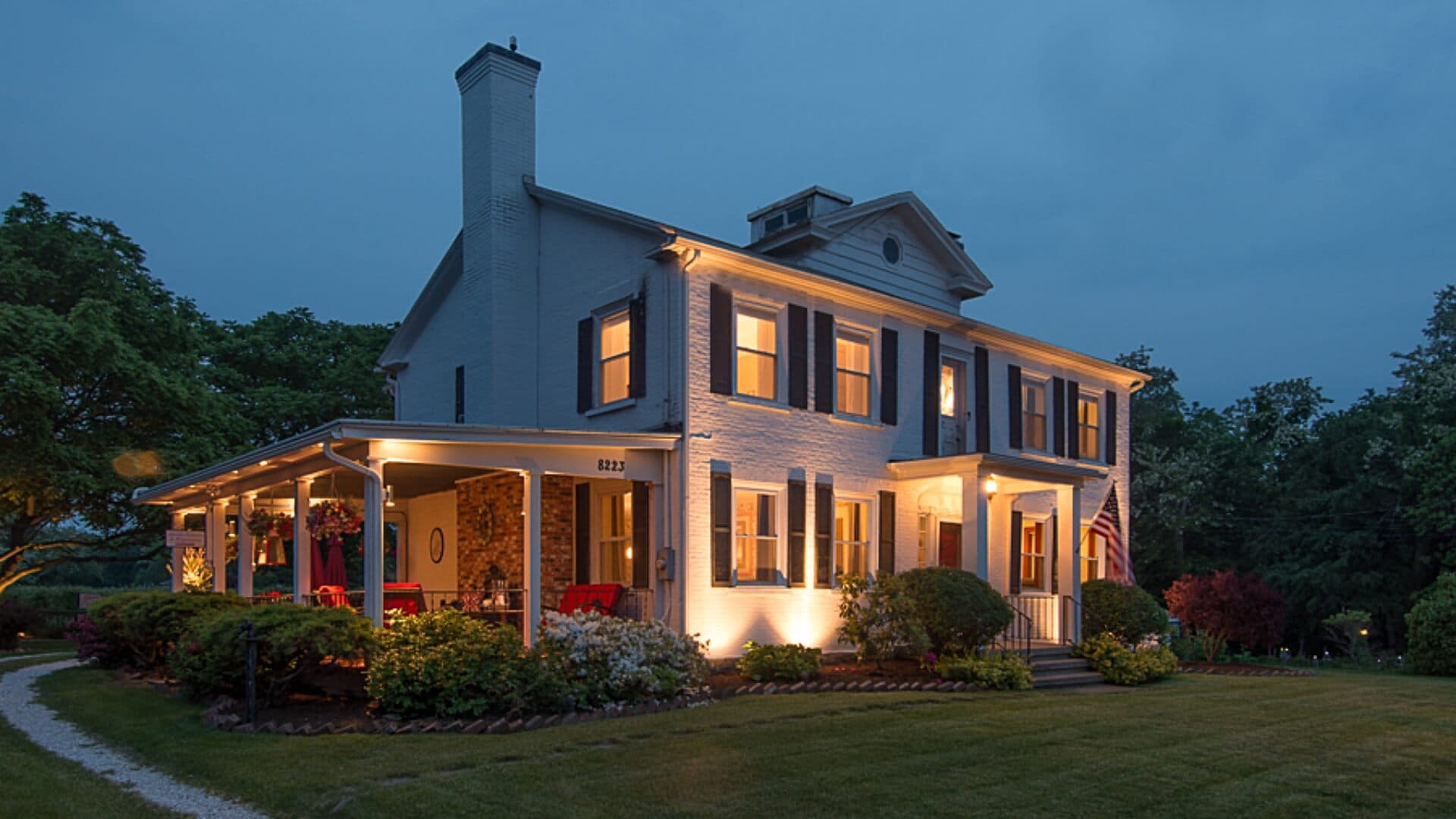A two-story white house with a porch, warmly lit at dusk, surrounded by greenery.